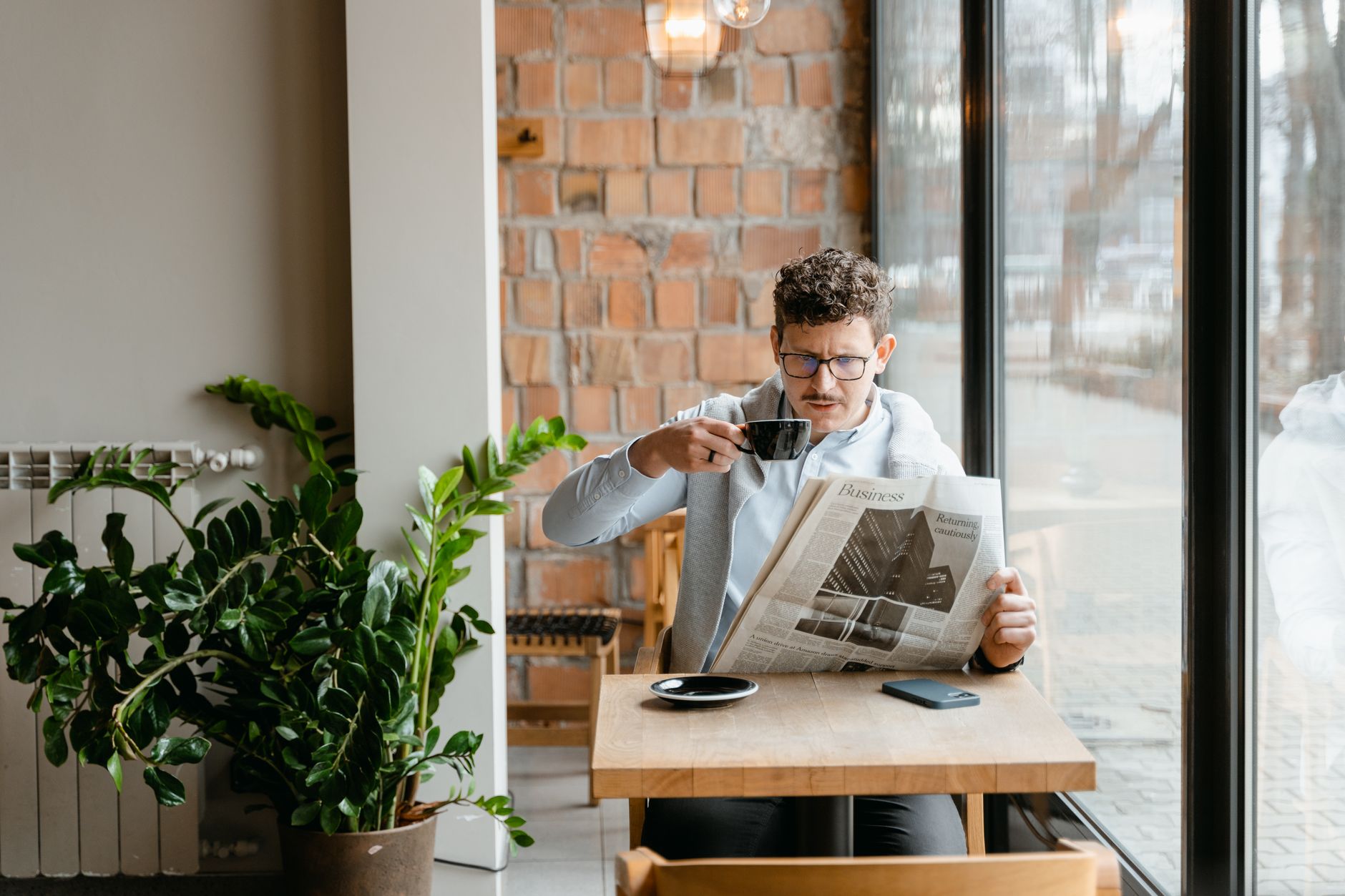 Man sitting indoors with coffee and newspaper, surrounded by plants by the window. — Warum neue Spieler GGBet Casino ausprobi